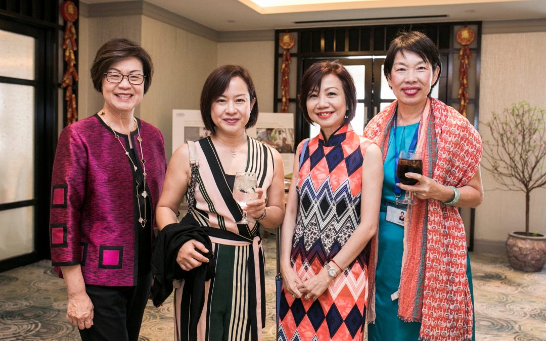 Four women standing together in a room with a table, engaged in a conversation and displaying a sense of camaraderie.